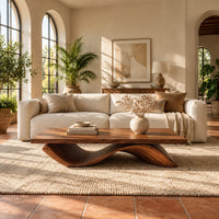 Rectangular coffee table with a flat plank top on a single rolling wave base in a bright living room with arched windows and plants