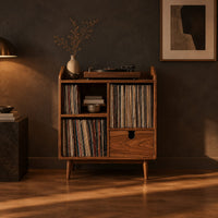 Wood record player stand on tapered legs with open cubbies and a drawer in a dark living room at golden hour