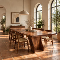 Rectangular dining table with a thick slab top on a wide hourglass pedestal in a bright dining room with arched windows and plants