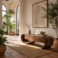 Looping wooden bench with a ribbon shaped frame and two drawers in a bright entryway with arched windows and plants