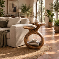 Side table with a looped ring base and two scalloped dish tops in a bright living room with arched windows and plants