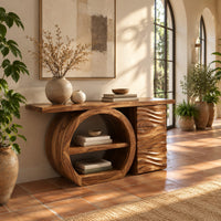 Console table with a large circular open shelf frame beside a wave carved cabinet in a bright entryway with arched windows and plants