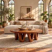 Round coffee table with a wide flat top on chunky v shaped legs in a bright living room with arched windows and plants