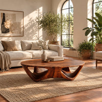 Round coffee table with a flared sculpted base and scooped arch openings in a bright living room with arched windows and plants