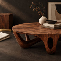 Corner detail of round coffee table with arched sculpted legs and oval openings beside wood, brass and linen samples