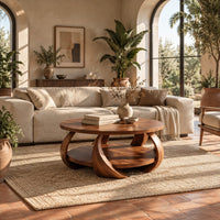 Round coffee table with crescent shaped legs and a round lower shelf in a bright living room with arched windows and plants