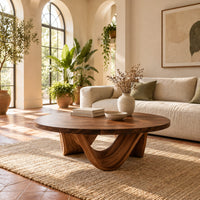 Round coffee table with a low swooping arch base in a bright living room with arched windows and plants