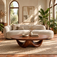 Round coffee table with a thick top on a sculpted base with two arches in a bright living room with arched windows and plants