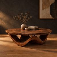 Round coffee table with a wavy ribbon base and swirled wood grain in a dark studio against a charcoal plaster wall
