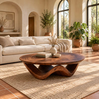 Round coffee table with a wavy ribbon base and swirled wood grain in a bright living room with arched windows and plants