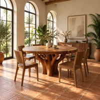 Round dining table with a carved tree trunk base and a grained top in a bright dining room with arched windows and plants