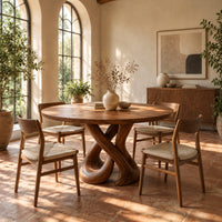 Round dining table on a sculpted base of two crossing wooden loops in a bright dining room with arched windows and plants