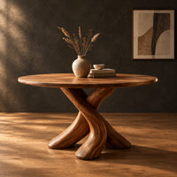 Circular dining table with two crossed branch shaped legs and wide curved feet in a dark studio against a charcoal plaster wall