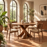 Circular dining table with two crossed branch shaped legs and wide curved feet in a bright dining room with arched windows and plants