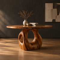 Circular wooden dining table on a sculpted pedestal with an open oval center in a dark studio against a charcoal plaster wall