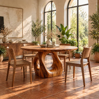 Circular wooden dining table on a sculpted pedestal with an open oval center in a bright dining room with arched windows and plants