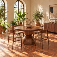 Round dining table with a radiating grain top and a sculpted hourglass base in a bright dining room with arched windows and plants