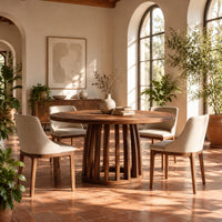 Circular dining table with a slatted drum pedestal base in a bright dining room with arched windows and plants