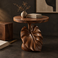 Corner detail of round side table on a pedestal carved like a large leaf beside wood, brass and linen samples