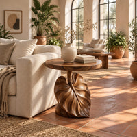 Round side table on a pedestal carved like a large leaf in a bright living room with arched windows and plants