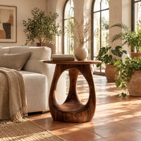 Round side table with an open teardrop pedestal and a flared foot in a bright living room with arched windows and plants