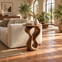 Round wood side table on a twisted figure eight pedestal in a bright living room with arched windows and plants