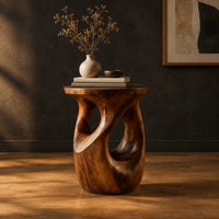 Round side table with a vase shaped base and twisting cutouts in a dark studio against a charcoal plaster wall