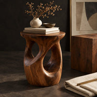 Corner detail of round side table with a vase shaped base and twisting cutouts beside wood, brass and linen samples