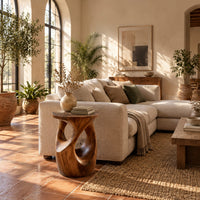 Round side table with a vase shaped base and twisting cutouts in a bright living room with arched windows and plants