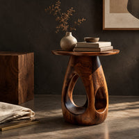 Corner detail of round side table with teardrop cutouts in a pear shaped pedestal beside wood, brass and linen samples