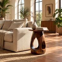 Round side table with teardrop cutouts in a pear shaped pedestal in a bright living room with arched windows and plants