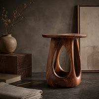 Corner detail of side table with a round wood top and a bulging base with teardrop cutouts beside wood, brass and linen samples