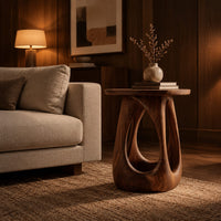 Side table with a round wood top and a bulging base with teardrop cutouts in an evening living room with walnut wall paneling