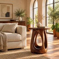 Side table with a round wood top and a bulging base with teardrop cutouts in a bright living room with arched windows and plants