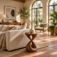 Side table with a circular top and a twisted figure eight pedestal base in a bright living room with arched windows and plants