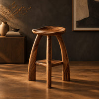 Saddle seat bar stool with curved legs and a low square shelf in a dark studio against a charcoal plaster wall
