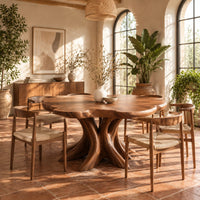 Dining table with a scalloped edge top and a flared branching pedestal in a bright dining room with arched windows and plants