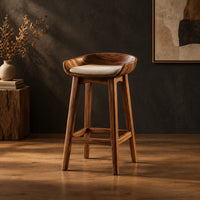 Bar stool with a scooped wooden seat and a cream cushion in a dark studio against a charcoal plaster wall