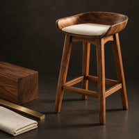 Corner detail of bar stool with a scooped wooden seat and a cream cushion beside wood, brass and linen samples
