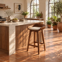 Bar stool with a scooped wooden seat and a cream cushion in a bright kitchen with arched windows and plants