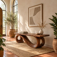 Wooden bench with a thick plank seat on a flowing scroll shaped base in a bright entryway with arched windows and plants
