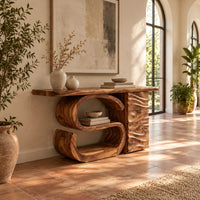 Console table with a large scroll base and a wave carved side column in a bright entryway with arched windows and plants