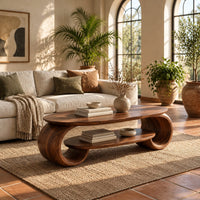 Oval coffee table with scrolled end supports and a lower oval shelf in a bright living room with arched windows and plants