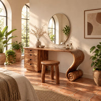Makeup vanity with four drawers and a sweeping top ending in a scroll leg in a bright bedroom with arched windows and plants