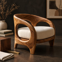 Corner detail of barrel shaped chair with open oval cutouts and a cream seat cushion beside wood, brass and linen samples