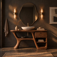Freeform vanity cabinet with a swooping leg and a small drawer above open cubbies in a dark bathroom at golden hour