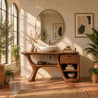 Freeform vanity cabinet with a swooping leg and a small drawer above open cubbies in a bright bathroom with arched windows and plants