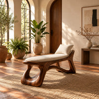Sculpted bench with a cushioned seat and open loop legs in a bright entryway with arched windows and plants