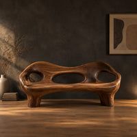 Sculpted wood bench with a low back and three oval openings in a dark studio against a charcoal plaster wall