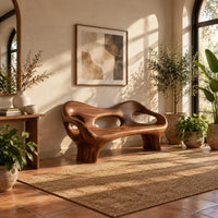 Sculpted wood bench with a low back and three oval openings in a bright entryway with arched windows and plants